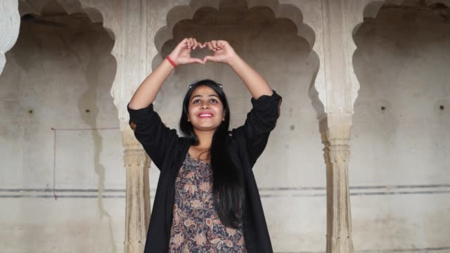 Happy-woman-walks-into-frame-and-admires-an-ancient-Indian-temple-moves-her-hands-over-her-head-looks-to-camera-makes-heart-sign-with-fingers-inside-building-interior-handheld-follows-smiles-fun-love