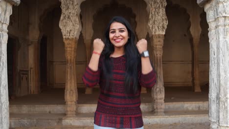 Gorgeous-young-woman-gets-good-news-wears-smart-watch-happy-thrilled-celebrate-fists-in-the-air-looks-at-camera-in-front-of-a-tradition-Indian-architectural-facade-Handheld-stabilized-mid-shot-learns
