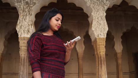 Handheld-Schuss-der-jungen-Frau-auf-ihrem-Handy-Handy-smart-kommunizieren,-Gespräche-Gespräche-Nachricht-Bildschirmtexte-berühren-Anteil-Netzwerk-Lächeln-vor-indischen-Architektur-Spalte-Bogen-Kulisse-hautnah