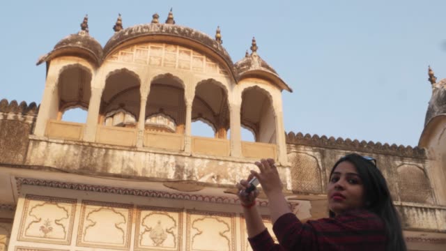 Handheld-shot-of-tourist-woman-in-front-of-Indian-Hindu-temple-taking-selfie--photo-video-on-her-mobile-smart-phone-happy-share-facade-architecture-ancient-holy-religious-fort-castle-palace-Rajasthani