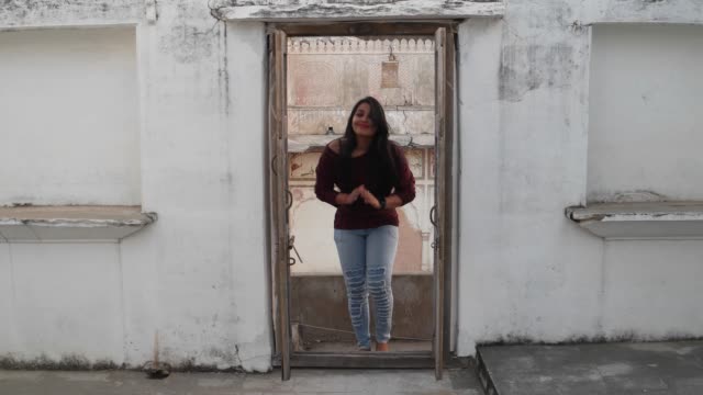 Young-woman-enters-and-exits-an-old-building-doorway-with-folded-joined-hands-to-namaste,-a-traditional-Indian-greeting-for-respect-laughs-smiles-points-hands-youngster-waves-jovial-elders-handheld