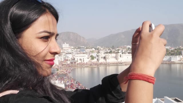 Handheld-Frau-nimmt-Selfie-Foto-Video-mit-ihrem-mobilen-Handy-Gerät-von-einem-Panorama-Aussichtspunkt-Blick-des-Heiligen-Pushkar-See,-eine-heilige-Stadt-für-Hindu-Pilger-Mela-Festival-Baden-Ghats-Tempel