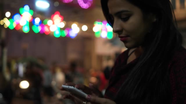 Young-Indian-woman-on-a-touch-screen-smart-mobile-phone-texts-messages-types-shares-photo-video-calls-in-front-of-a-festival-colorful-bright-lights-out-of-focus-in-the-background-celebration-mela-love