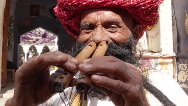 Hand-held-CU-Rajasthani-elderly-male-plays-the-flute-with-his-nose-in-front-of-a-painted-temple-archway,-with-big-moustache-wearing-traditional-attire