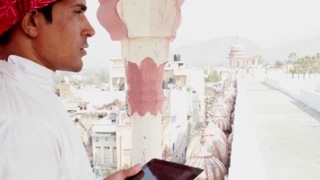 Indischer-Mann-hektisch-auf-einem-Tablet-arbeiten-und-denken-auf-einem-Hausdach-in-traditioneller-Kleidung-in-Rajasthan,-Indien