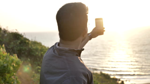 Ein-glücklicher-junger-Mann-oder-Reisenden-unter-ein-Bild-eines-Sonnenuntergangs-auf-sein-Handy-oder-Smartphone-der-untergehenden-Sonne.