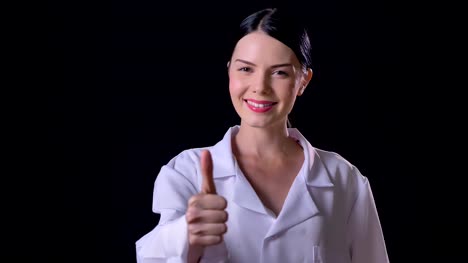 Confident-young-female-scientist-in-white-coat-showing-thumbs-up-into-camera-and-smiling,-isolated-on-black-background