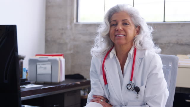 Senior-female-doctor-sitting-at-desk-in-an-office,-smiling