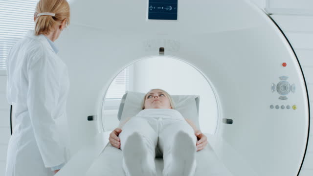 En-el-laboratorio-médico-paciente-femenino-acostado-en-una-cama-de-exploración-de-MRI-o-CT-somete-a-análisis-el-procedimiento-bajo-la-supervisión-de-un-radiólogo-profesional.-Paciente-se-mueve-a-través-de-la-máquina-mientras-que-explora.