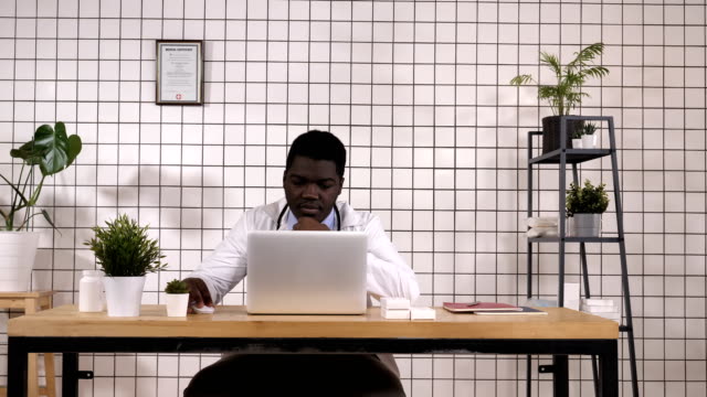 Médico-joven-afroamericano-en-el-trabajo,-con-ordenador-portátil