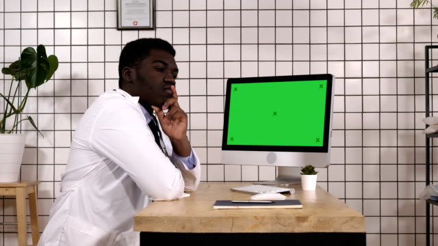 Männlichen-schönen-afrikanischen-Arzt-neben-seinen-Computer-denken.-Weißes-Display