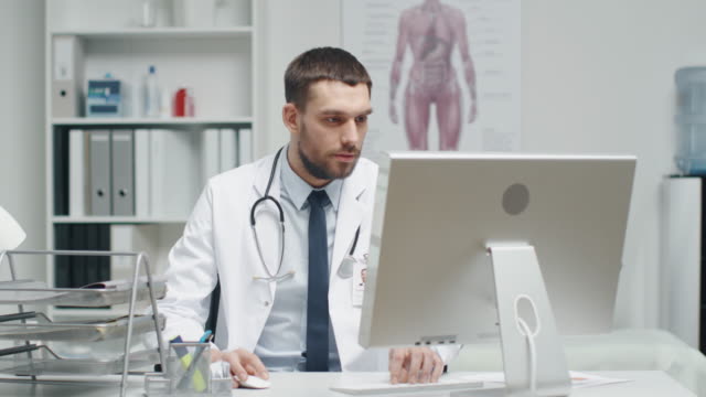 Male-Doctor-is-Working-at-His-Desk.-He-Interrupts-His-Work,-Folds-His-Hands-and-Smiles-at-the-Camera.