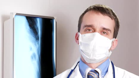 Ein-junger-Arzt-in-eine-Gesichtsmaske-befasst-sich-mit-der-Kamera-und-lächelt.-Vor-dem-Hintergrund-hängen-Röntgenaufnahmen-des-Patienten.-Hemd-mit-Krawatte-und-ein-Stethoskop-auf-den-Hals