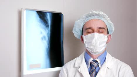 Ein-junger-Arzt-in-eine-Gesichtsmaske-befasst-sich-mit-der-Kamera-und-lächelt.-Schutzhelm-auf-dem-Kopf.-Vor-dem-Hintergrund-hängen-Röntgenaufnahmen-des-Patienten.-Hemd-mit-Krawatte-und-ein-Stethoskop-auf-den-Hals