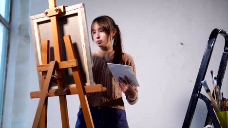 Young-woman-paints-oil-paints-on-canvas-in-art-space-with-large-windows