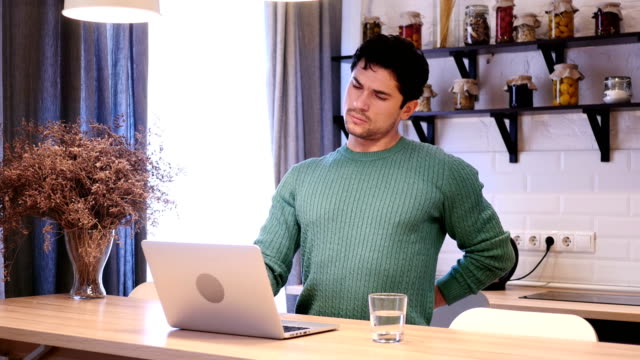 Young-Man-with-Back-Pain-Working-on-Laptop--in-Kitchen