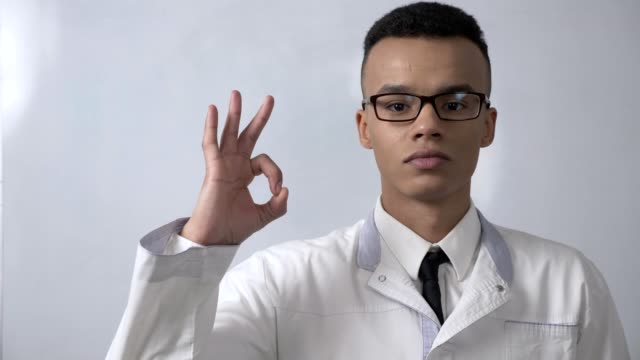 Young-successful-African-scientist-in-a-white-coat-and-glasses-showing-ok-sign,-looks-at-the-camera,-portrait-concept.-60-fps