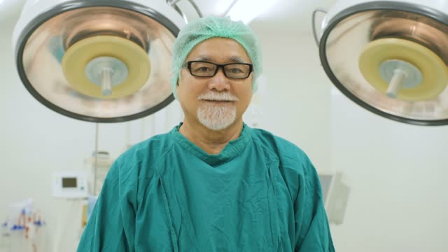 Portrait-of-senior-male-surgeon-wearing-full-surgical-scrubs-smiling-camera-in-operating-theater-at-the-hospital.