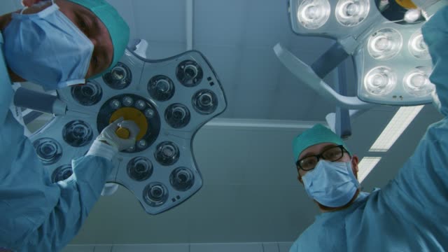 Low-Angle-Shot-POV-Patient-View:-Two-Professional-Surgeons-Turning-on-Surgery-Lights-while-Bending-over-Patient.
