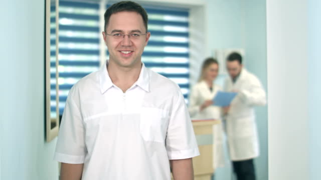 Lächelnde-männlichen-Arzt-in-Gläsern,-Blick-in-die-Kamera-während-des-medizinischen-Personals-arbeiten-auf-dem-Hintergrund