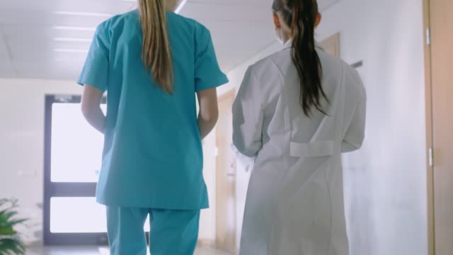 En-el-Hospital,-vista-posterior-tiro-de-enfermera-y-Doctor-caminando-por-el-pasillo.-Personal-del-hospital-trabajando.
