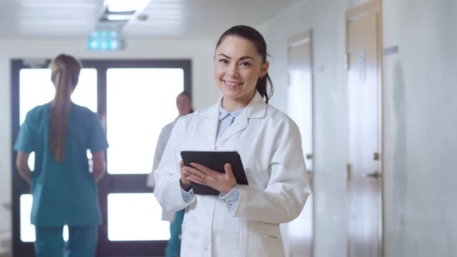 Beautiful-Young-Female-Doctor-Stands-in-the-Hospital-Hallway,-Uses-Tablet-Computer-and-Charmingly-Smiles.-Professional-People-at-Work.