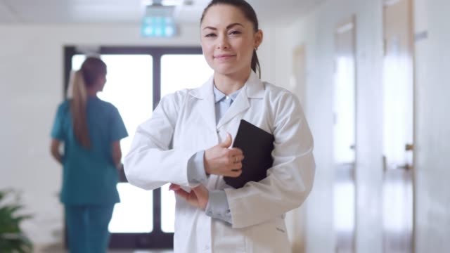 Beautiful-Young-Female-Doctor-Walks-Through-the-Hospital-Hallway,-She-Stops,-Crosses-Arms-and-Charmingly-Smiles.-Professional-People-at-Work.
