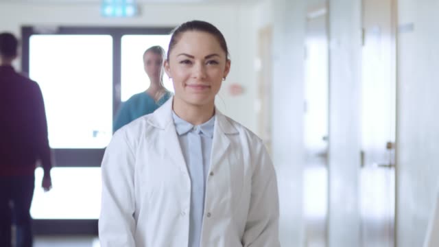 Beautiful-Young-Female-Doctor-Walks-Through-the-Hospital-Hallway,-She-Stops,-Crosses-Arms-and-Charmingly-Smiles.-Professional-People-at-Work.