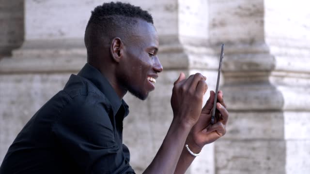 lächelnde-junge-schwarze-afrikanische-verwendet-Tablet-sitzt-auf-der-Treppe-in-der-Stadt