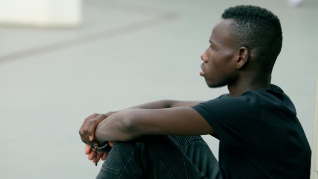 Perfil-de-triste-solitario-negro-africano-joven-sentado-en-el-suelo-al-aire-libre