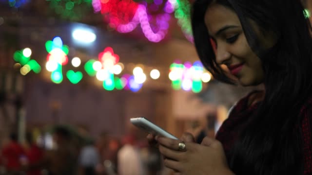 Young-Indian-woman-on-a-touch-screen-smart-mobile-phone-texts-messages-types-shares-photo-video-calls-in-front-of-a-festival-colorful-bright-lights-out-of-focus-in-the-background-celebration-mela-love