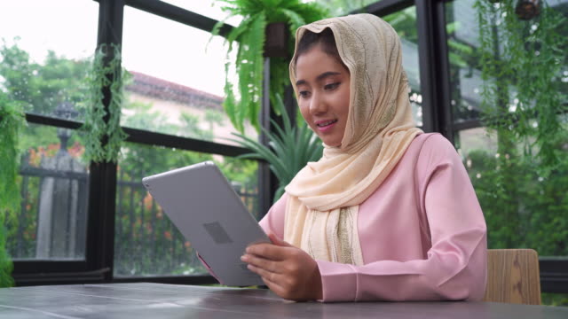 Hermosa-joven-sonriente-a-mujer-musulmana-asiática-utilizando-tablet-sentado-en-la-sala-de-estar-en-casa.-Disfrutando-en-casa.