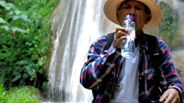 Close-Up-Senior-man-sitting-enjoying-by-a-waterfall-and-drink-Water.-Travel-Lifestyle-vacations-into-the-wild-wearing-cozy-shirt,hat-with-backpack.-Video-Slow-motion