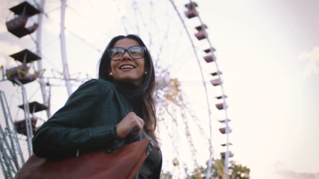 Mujer-joven-con-estilo-en-gafas-sonriendo-alegremente-delante-de-una-noria-en-el-parque-de-atracciones