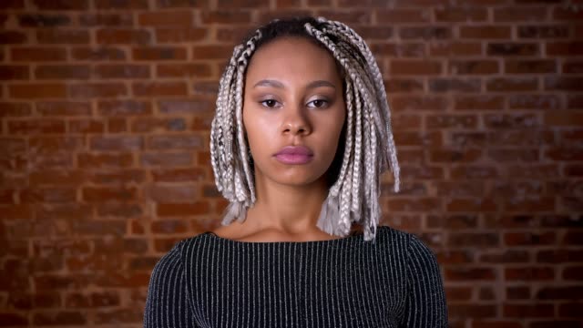 Young-African-serious-girl-with-dreadlocks-girl-looking-at-camera,-closing-eyes,-Brick-wall-in-the-background.
