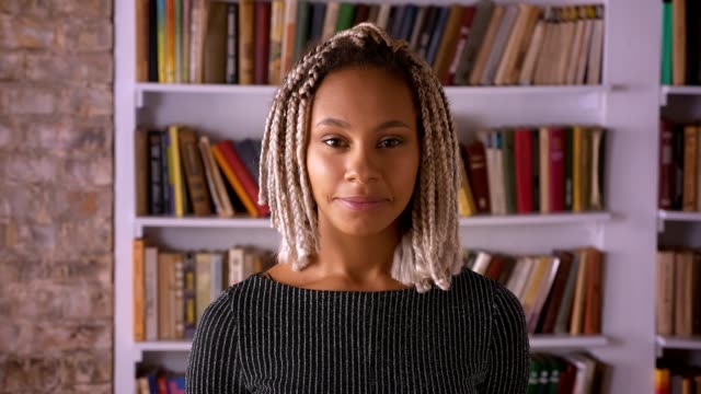 Young-African-girl-with-dreadlocks-smiling-and-looking-at-camera,-Book-shelves-in-the-background