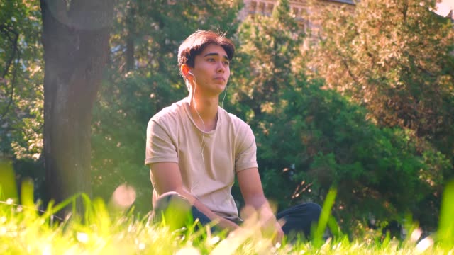 Dreaming-asian-boy-is-hanging-out-alone-in-park,-sitting-on-the-grass-in-sunlights,-fresh-spring-atmosphere