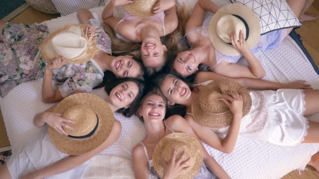 joyful-girlfriends-in-pajamas-lie-on-bed-with-straw-hats-on-faces-and-then-take-off-them-and-smile-into-camera-during-hen-party