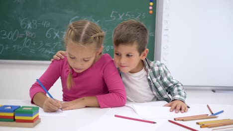 Schule-Liebe-Klassenkameraden-während-Lektion-am-Tisch-zu-schreiben,-und-dann-in-die-Kamera-schauen-und-Lächeln-auf-Hintergrund-von-blackboard