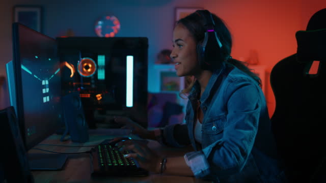 Porträt-von-einem-hübschen-und-aufgeregt-schwarz-Gamer-Mädchen-Kopfhörer-spielen-Ego-Shooter-Online-Video-Spiel-auf-ihrem-Computer.-Sie-dreht-sich-und-lächelt-in-die-Kamera.-Haus-ist-mit-Neonröhren-beleuchtet.