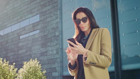 Elegante-Geschäftsfrau-nutzt-Smartphone-zu-handeln-in-der-Wirtschaft-während-stehen-außen-modernen-Glasbau.-Schöne-stilvolle-Frau-tragen-Fell-und-eine-dunkle-Brille-geht-im-urbanen-Umfeld-der-modernen-Stadt.