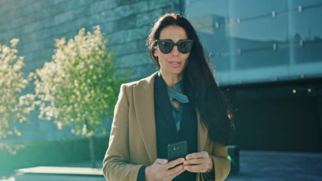 Elegante-empresaria-usa-Smartphone-para-realizar-negocios-mientras-camina-por-el-edificio-de-cristal-moderno.-Hermosa-mujer-elegante-vestida-con-abrigo-y-gafas-oscuras-paseos-en-entorno-urbano-de-la-ciudad-moderna.
