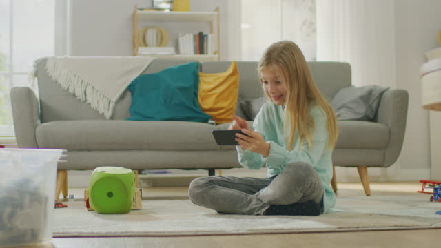 Chica-linda-inteligente-sentado-sobre-una-alfombra-jugando-en-juego-de-Video-en-su-teléfono-inteligente,-tiene-y-usa-teléfono-móvil-en-modo-apaisado-Horizontal.-Niño-tiene-diversión-jugando-videojuegos-en-la-sala-de-estar-soleado.