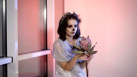 Close-up-of-a-young-girl-with-creative-scary-halloween-make.-in-appearance-dead-bride-sitting-on-the-bench-near-the-window-the-flower-in-hands-and-looking-at-the-camera