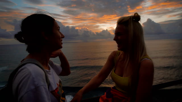 Zwei-glückliche-weibliche-Reisende-reden-und-lachen-bei-Sonnenuntergang-vor-dem-Hintergrund-des-Meeres.-Blonde-und-Brünette-Klappern-und-lächelnd-winkt-Ozean-hinter-ihnen-zu-betrachten