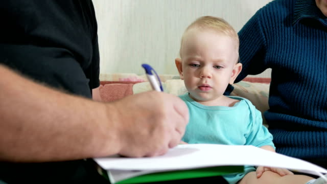 Attractive-baby-boy-draws-a-pen-with-his-grandparents-home-on-the-couch.-The-boy-stares-at-the-animals-that-drew-grandfather.-The-concept-of-different-generations