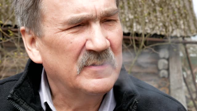 Un-anciano-con-un-bigote-fuma-en-la-calle-cerca-de-la-vieja-casa.-Vestido-con-un-suéter-negro