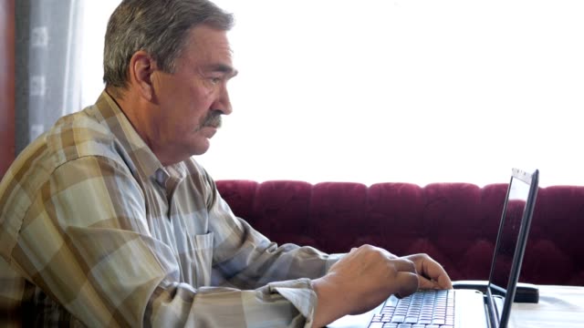 An-elderly-man-with-a-mustache-sits-behind-a-laptop-and-solves-problems.-He-looks-seriously-at-the-monitor