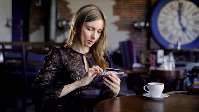 Lady-kaufen-online-mit-einer-Kreditkarte-und-Smartphone-sitzen-im-Restaurant-mit-Menschen-im-Hintergrund.-Frau-im-schwarzen-Kleid-mit-modernen-Handy-für-bargeldloses-Bezahlen-und-Einkäufen-im-Internet
