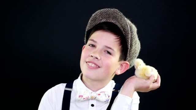 Portrait,-a-pretty-boy-in-a-cap-and-suspenders-plays-with-a-small-yellow-duckling.-Studio-video-with-thematic-decoration